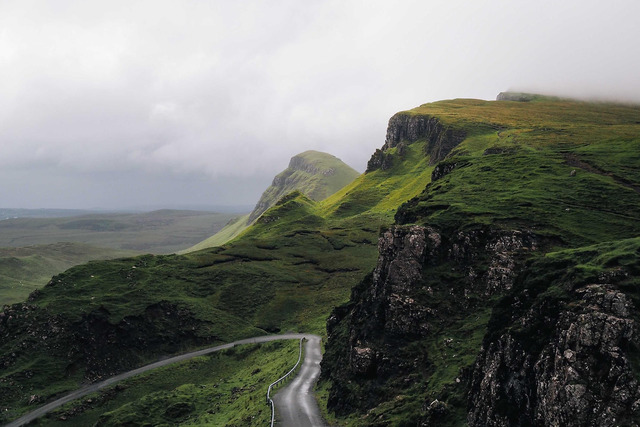 A narrow paved road with a guardrail curves through a green valley between steep grassy cliffs and a conical mountain peak under thick gray overcast clouds, with low mist over the upper right ridge.