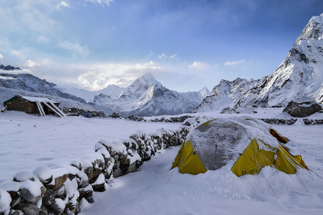 A snow-covered high-altitude campsite features a yellow and grey dome tent and a smaller second tent, set against snow-laden mountain peaks with a low stacked-stone wall running across the foreground.